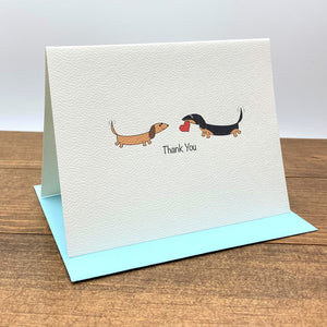 On the textured folded note card there are two dachshunds facing each other, one is brown and the other one is black and tan and holding a heart in its mouth.
