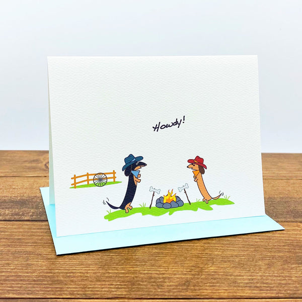 Two dachshunds wearing cowboy hats enjoying a bonfire on ranch.  The card message is "Howdy!"