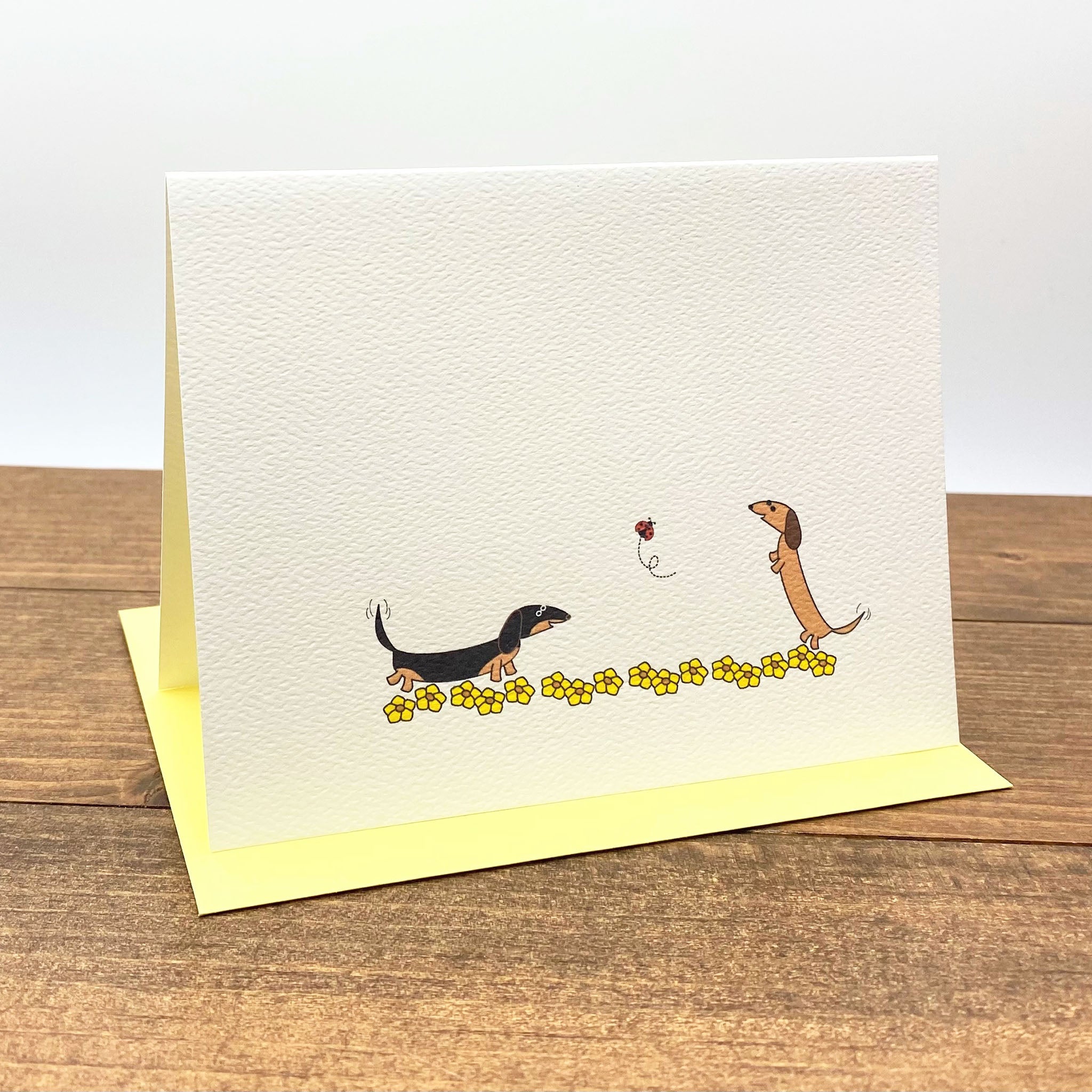 Note card with dachshunds playing with ladybug in field of yellow flowers.