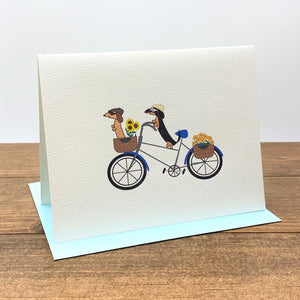 Two dachshunds riding a bicycle wearing riding caps and goggles,  with one dachshund in the front flower basket.