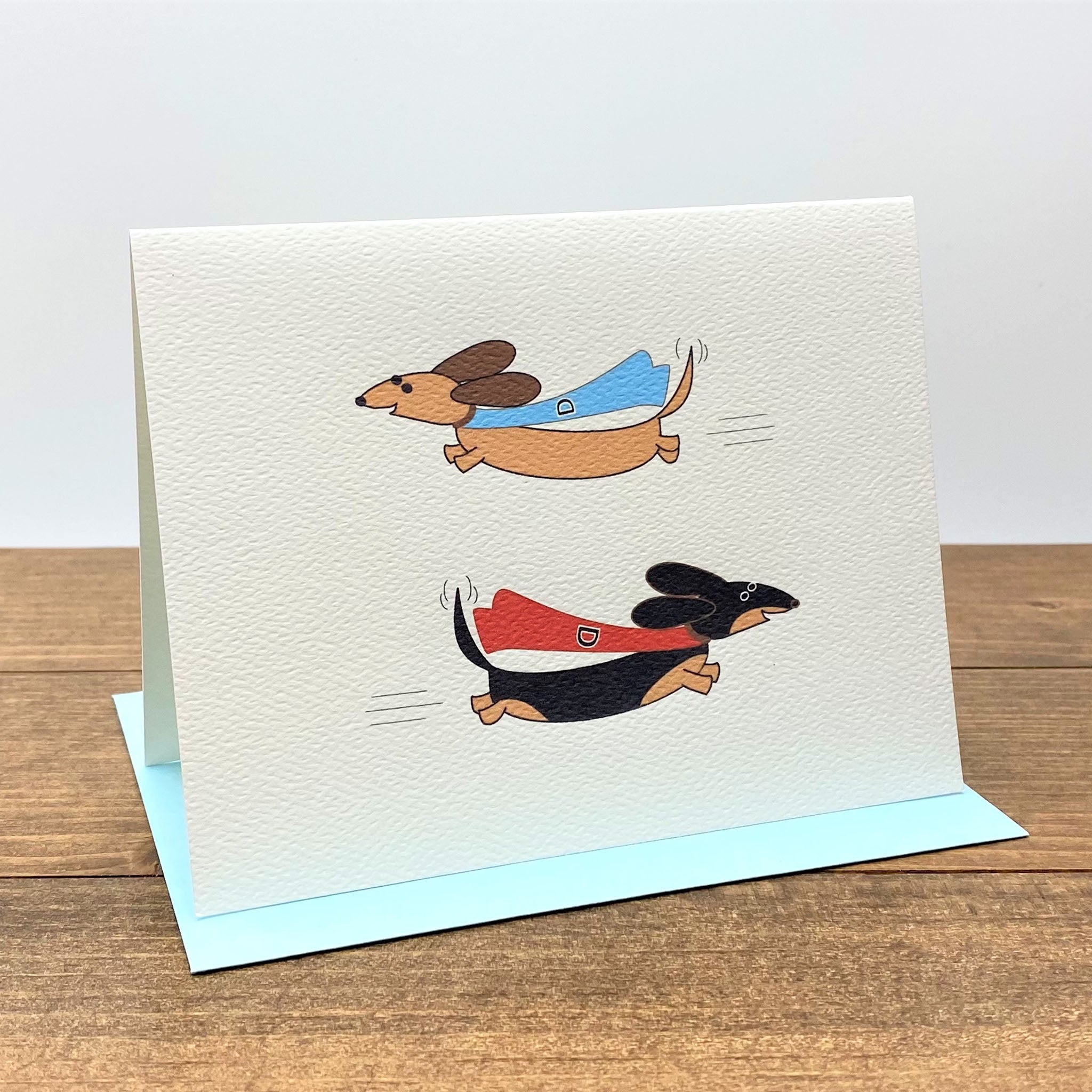Note card featuring two super dachshunds wearing capes and flying. 