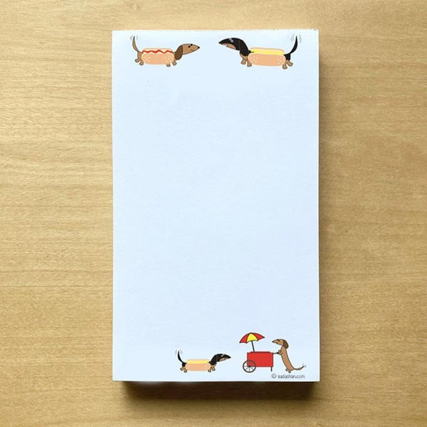 Two dachshunds dressed as hot dogs facing each other and a hot dog cart  in the bottom corner of this unlined notepad
