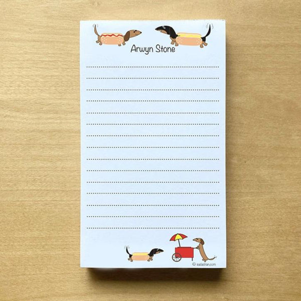 Two dachshunds dressed as hot dogs facing each other with personalized text below them and a hot dog cart  in the bottom corner of this lined notepad