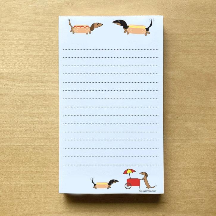 Two dachshunds dressed as hot dogs facing each other and a hot dog cart  in the bottom corner of this lined notepad