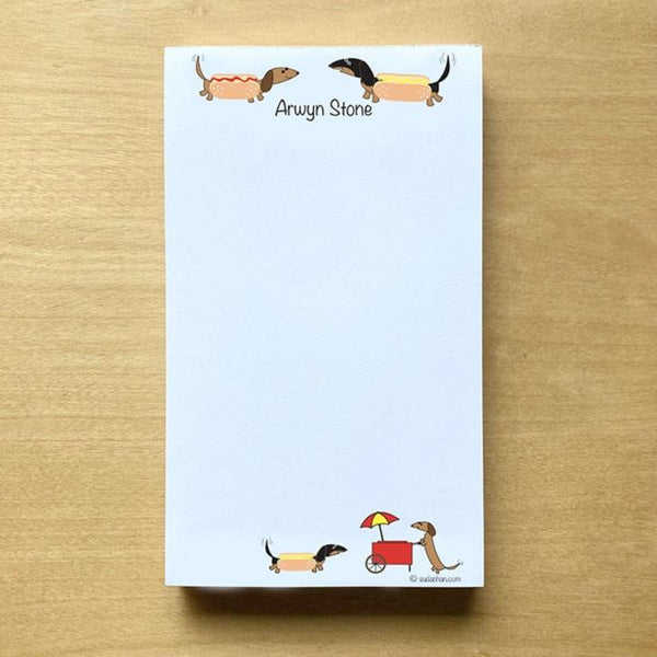 Two dachshunds dressed as hot dogs facing each other with personalized text below them and a hot dog cart  in the bottom corner of this unlined notepad