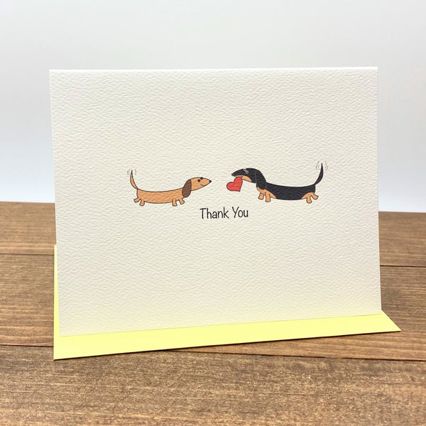 On the folded note card there are two dachshunds facing each other, one is brown and the other one is black and tan and holding a heart in its mouth.  The card  message is Thank You.