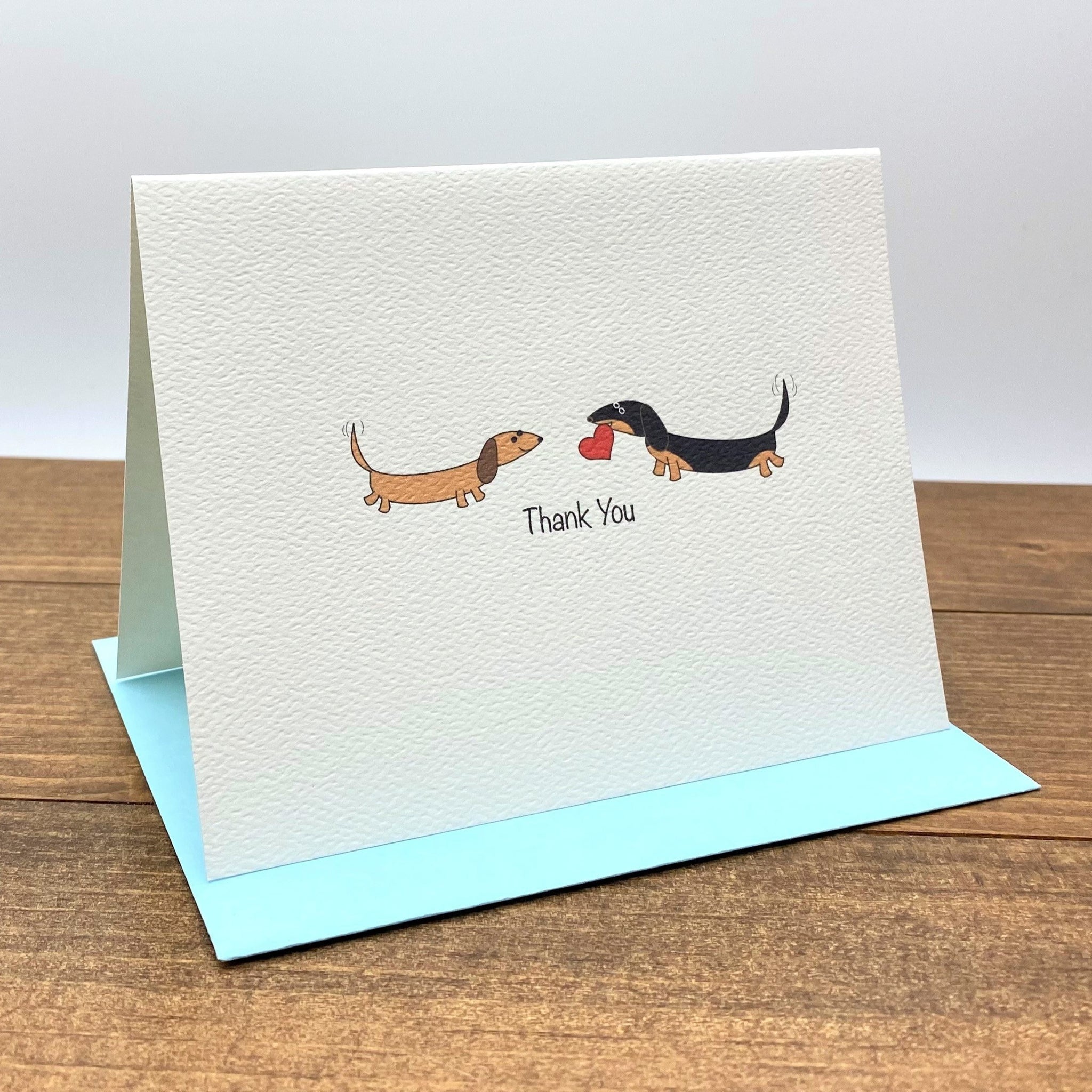 On the textured folded note card there are two dachshunds facing each other, one is brown and the other one is black and tan and holding a heart in its mouth.