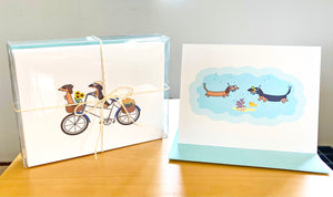 Traveling dachshunds assorted card set in clear stationery box tied with yellow twine, two card examples are two dachshund riding a bicycle and two dachshunds snorkeling under the sea