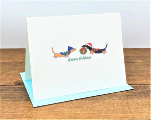 Two dachshunds wearing hats and scarves with wreath Happy Holidays Card