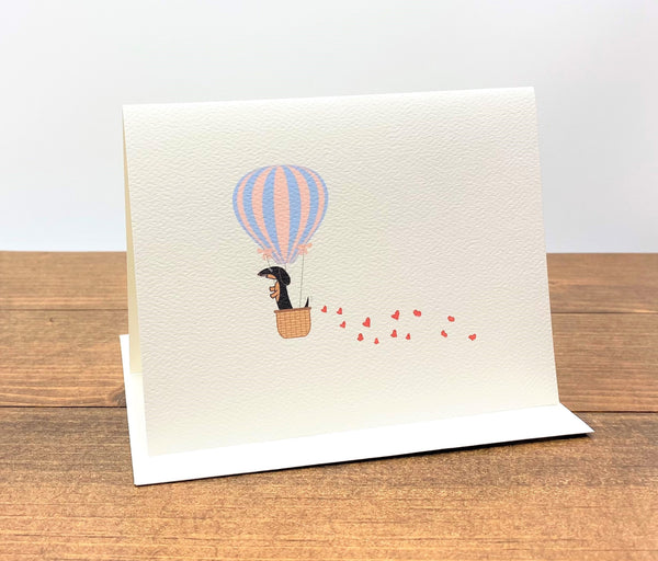 Note card with black and tan dachshund in hot air balloon with trail of hearts behind it.