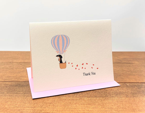 Thank you note card with black and tan dachshund in hot air balloon with trail of hearts behind it.